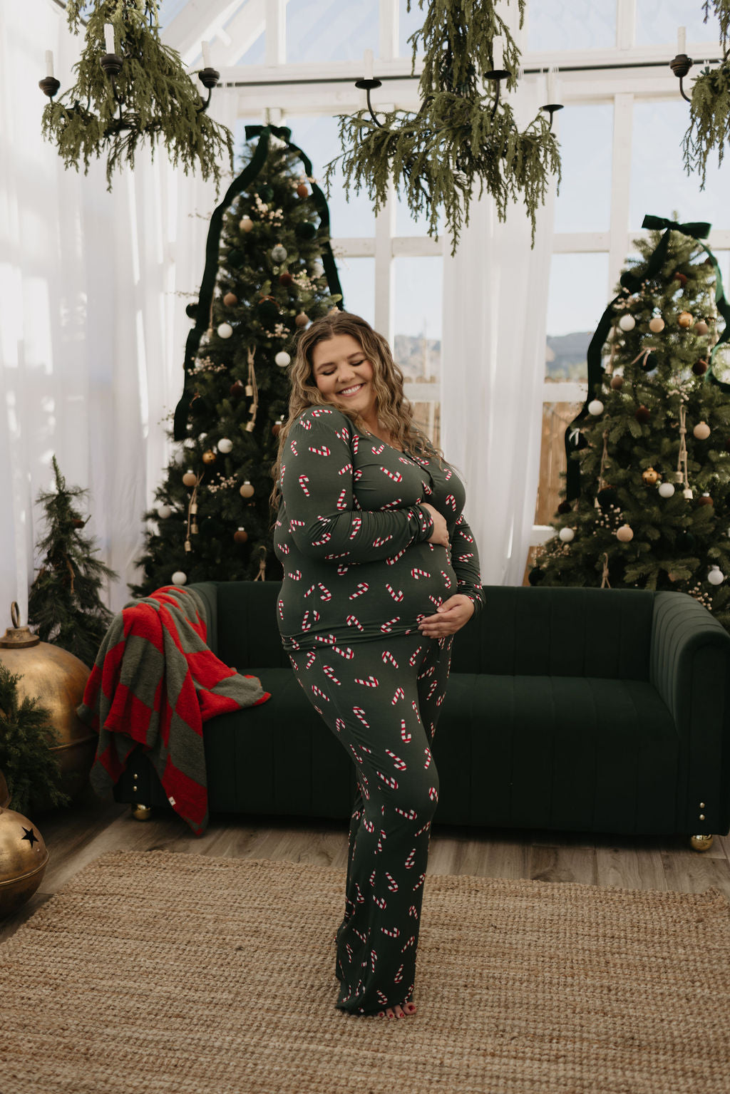 Wearing the Women's Bamboo Pajamas | Candy Cane Lane by forever french baby, a smiling person stands in a festive room. These hypo-allergenic dark green pajamas feature delightful candy cane patterns, ideal for breastfeeding moms during the holiday season. They pose in front of decorated Christmas trees, with ornaments and a plaid blanket adorning a dark green sofa in the background.