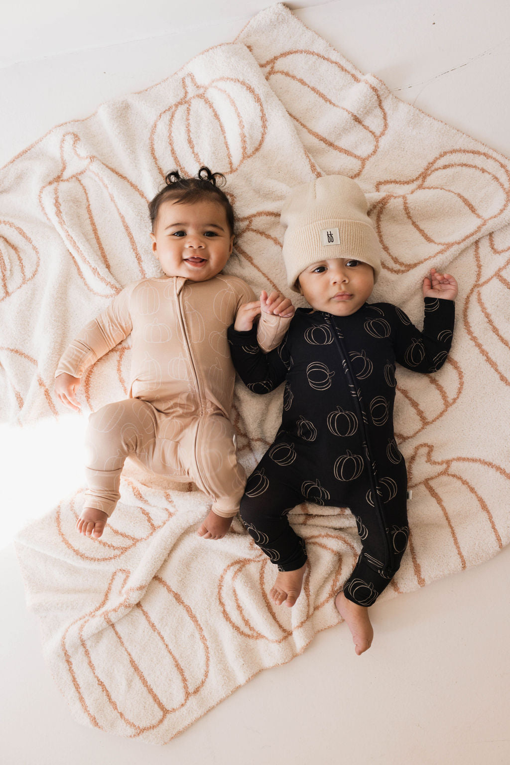 Two babies lie on a blanket with a pumpkin design. One wears a peach-colored onesie, and the other, in hypoallergenic Bamboo Zip Pajamas from forever french baby, sports a black onesie with a beanie. Both outfits feature a circular pattern. They are holding hands and looking up.