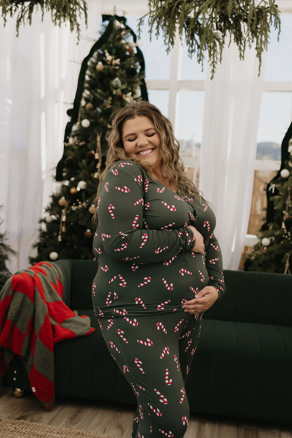 A smiling person wearing the Women's Bamboo Pajamas in the Candy Cane Lane pattern from forever french baby poses in front of Christmas trees indoors. The room, ideal for breastfeeding moms, is festively decorated with greenery and a red and green blanket draped over a couch, enhancing the cozy vibe.