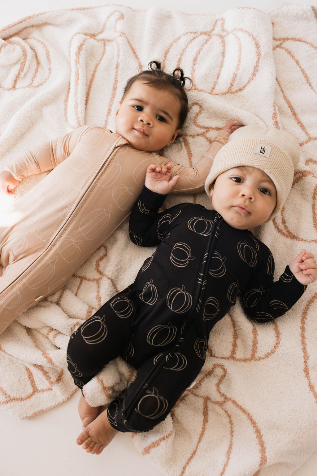 Two babies lying on a pumpkin-patterned blanket, surrounded by outlines of pumpkins. Both are wearing the Bamboo Zip Pajamas in Pumpkin Pie from forever french baby, one in beige and the other in black. The baby in black wears a cream knit hat while they both hold hands and look up.