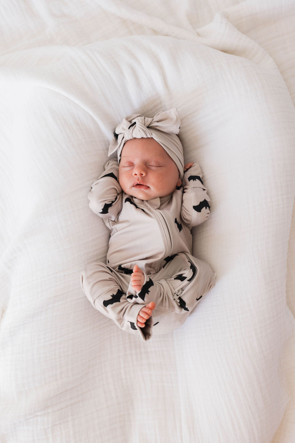 A newborn baby sleeps peacefully on a white blanket, dressed in a matching beige outfit with black animal prints. The baby wears an adjustable head wrap with a bow, specifically the "Bamboo Head Wrap | It's Bats!" from forever french baby. Their arms are raised beside their head. Designed for babies and toddlers, the outfit is made from soft bamboo material for ultimate comfort.