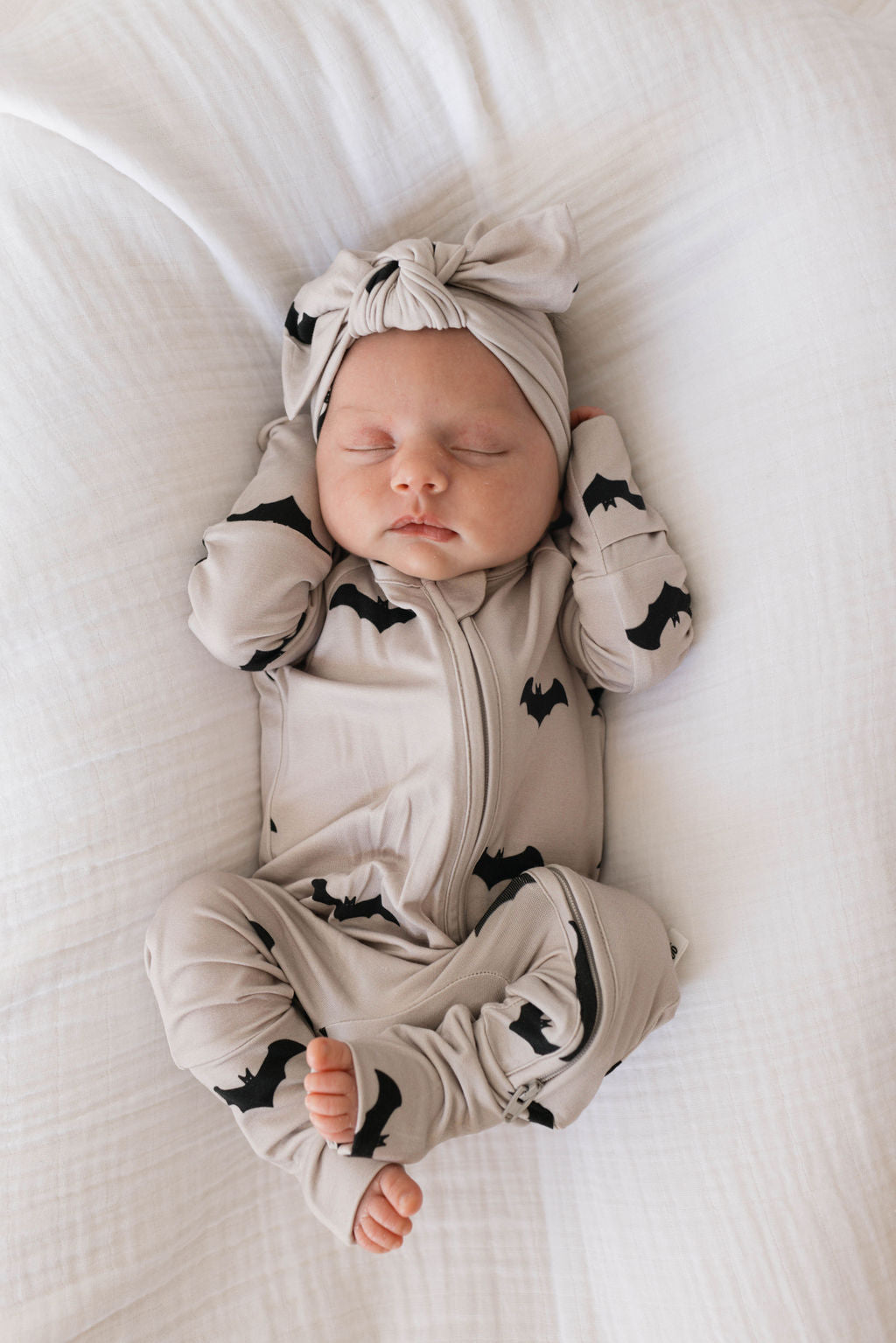 A newborn baby sleeps peacefully on a white blanket, wearing a light beige onesie adorned with black bat patterns. The baby also has an adjustable "Bamboo Head Wrap | It's Bats!" from the brand forever french baby, secured with a knot at the top. Their tiny hands are positioned near their head, embracing the comfort often preferred by babies and toddlers.