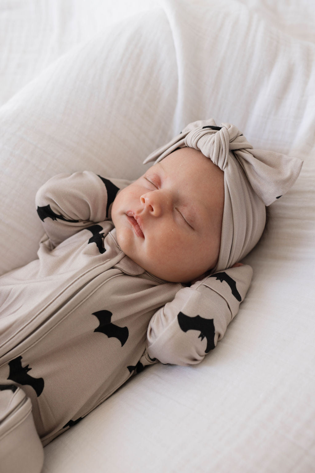 A sleeping baby dressed in a light beige onesie with black bat patterns lies on a white blanket. The cozy bamboo material ensures comfort, while the baby's matching light beige headband with a bow, the Bamboo Head Wrap | It's Bats! from forever french baby, is an adjustable head wrap. The baby's arms are bent with hands near its head.