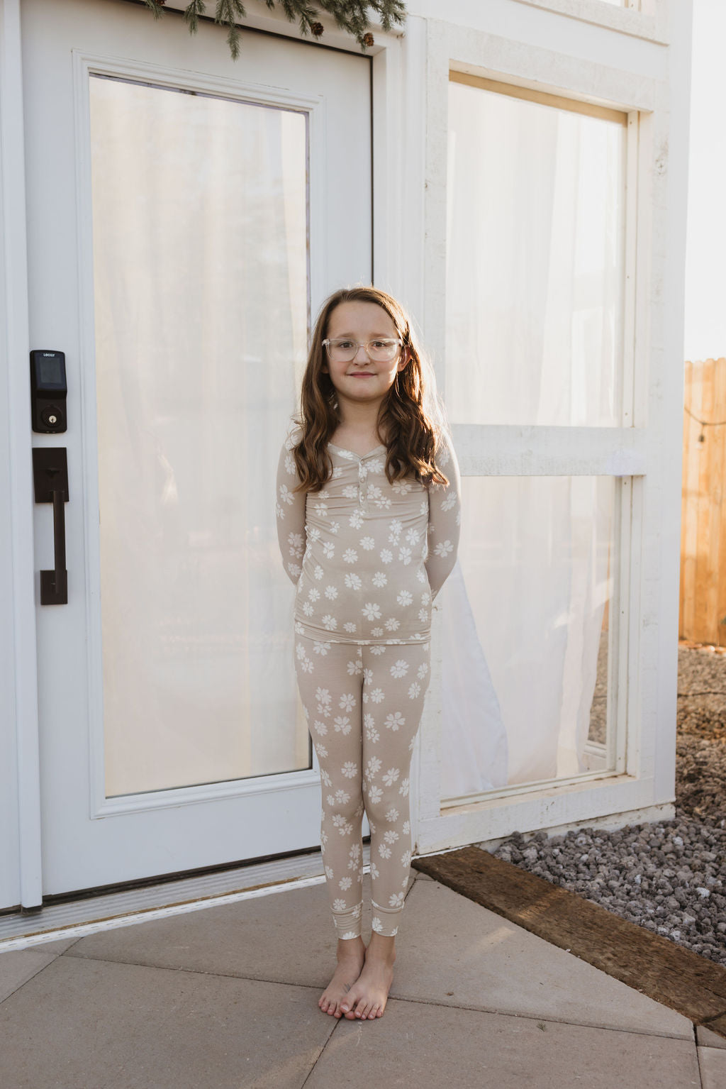 A young girl with glasses stands barefoot on a patio, wearing matching hypo-allergenic Bamboo Two Piece Pajamas from the Lazy Daisy collection by forever french baby. She is in front of a white glass door with a wooden frame, part of a building.