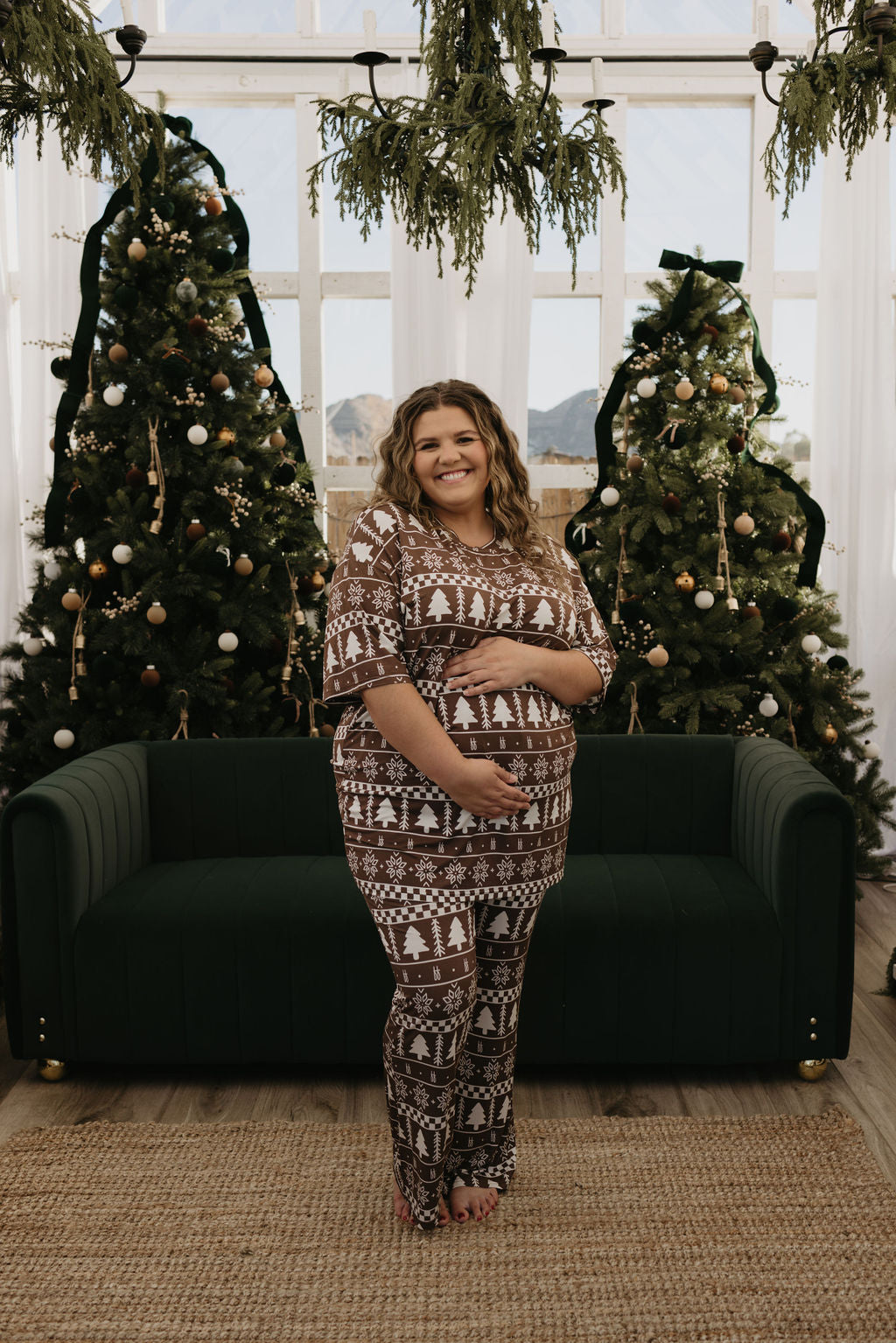 A smiling person stands in front of two Christmas trees, wearing the Short Sleeve Women's Bamboo Pajamas in the Forever Fair Isle pattern by forever french baby. The setting includes a large window and a dark green sofa, creating a warm and cheerful atmosphere with natural light illuminating the room.