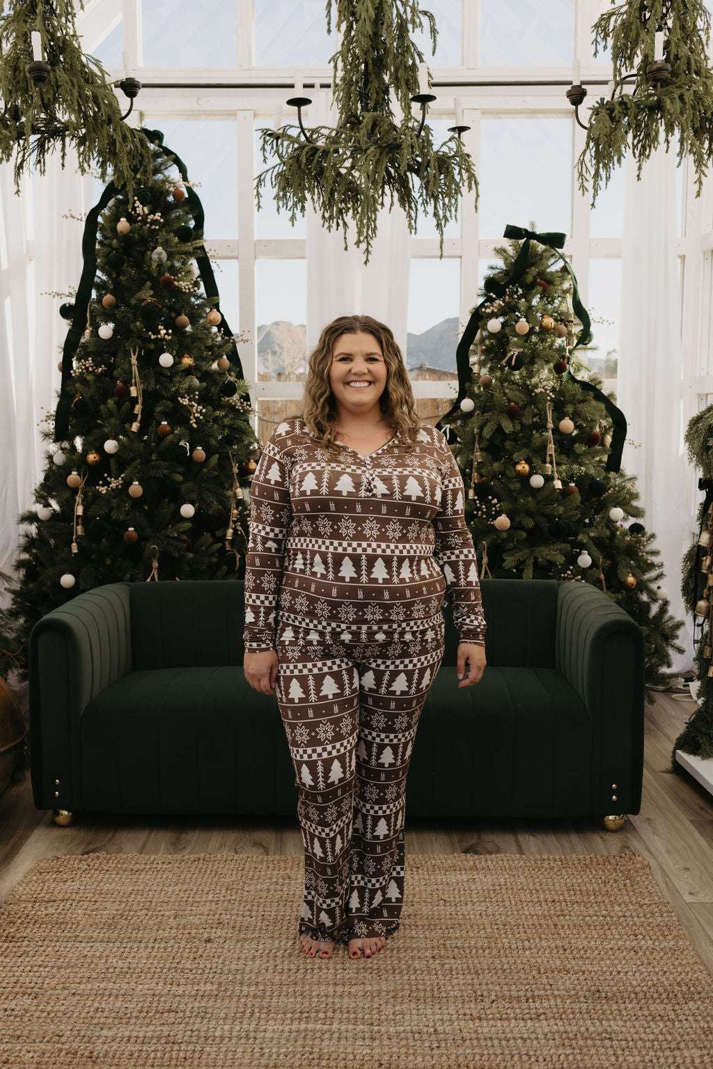 A person stands smiling in front of two decorated Christmas trees, dressed in cozy Women's Bamboo Pajamas designed with fair isle patterns from forever french baby. The setting is a warm indoor space with large windows and greenery.