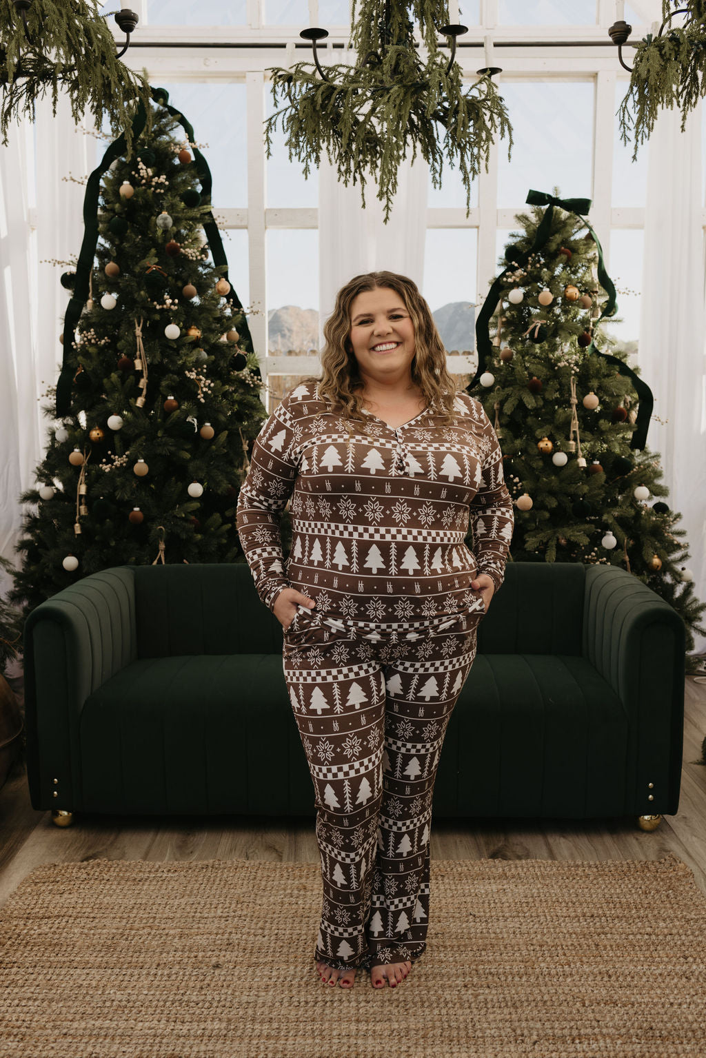 A person stands smiling in front of a green couch, wearing Women's Bamboo Pajamas | Forever Fair Isle from forever french baby, featuring tree and snowflake patterns. Behind them are two decorated Christmas trees, and soft light filters through large windows in the background, creating a cozy scene ideal for matching baby and toddler pajamas.