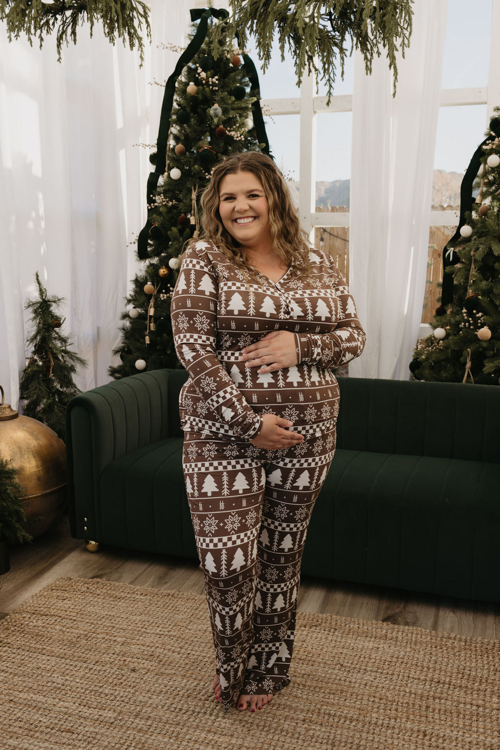 In a cozy room adorned with holiday decorations like a green sofa, Christmas trees, and garlands, someone is wearing Forever Fair Isle bamboo pajamas from forever french baby. They are smiling warmly and cradling their belly.