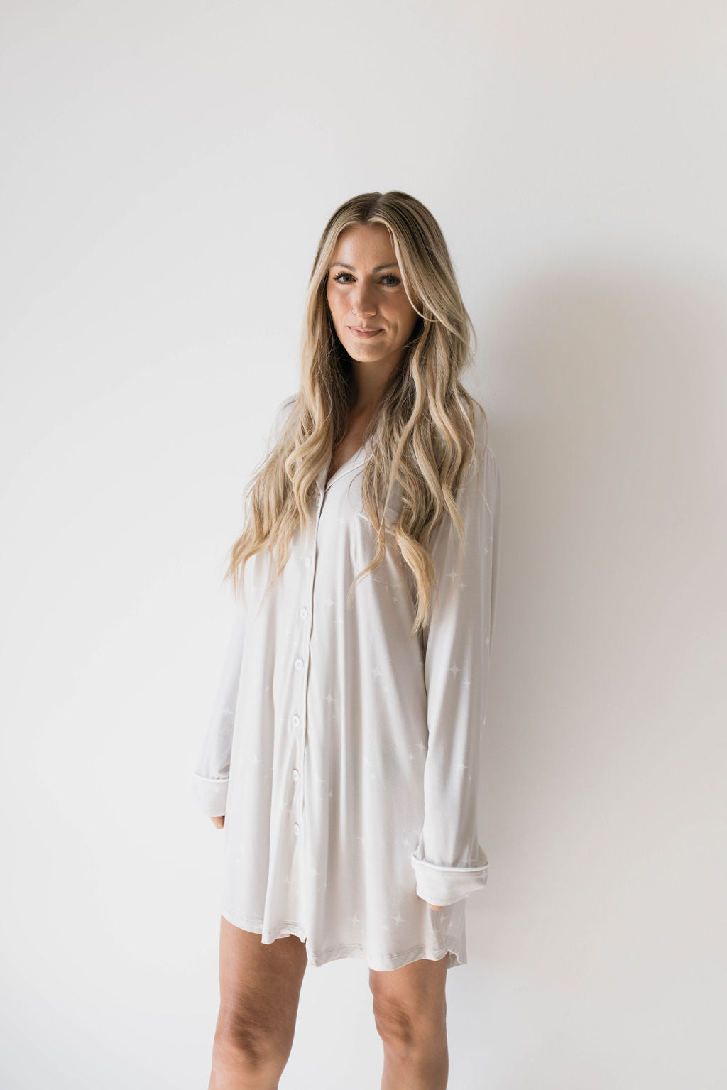 A person with long, wavy blond hair, wearing the Women's Bamboo Sleeping Dress from forever french baby, stands against a plain white background. They have a calm expression and are looking slightly to the side. The overall vibe of the image is minimalistic and serene.