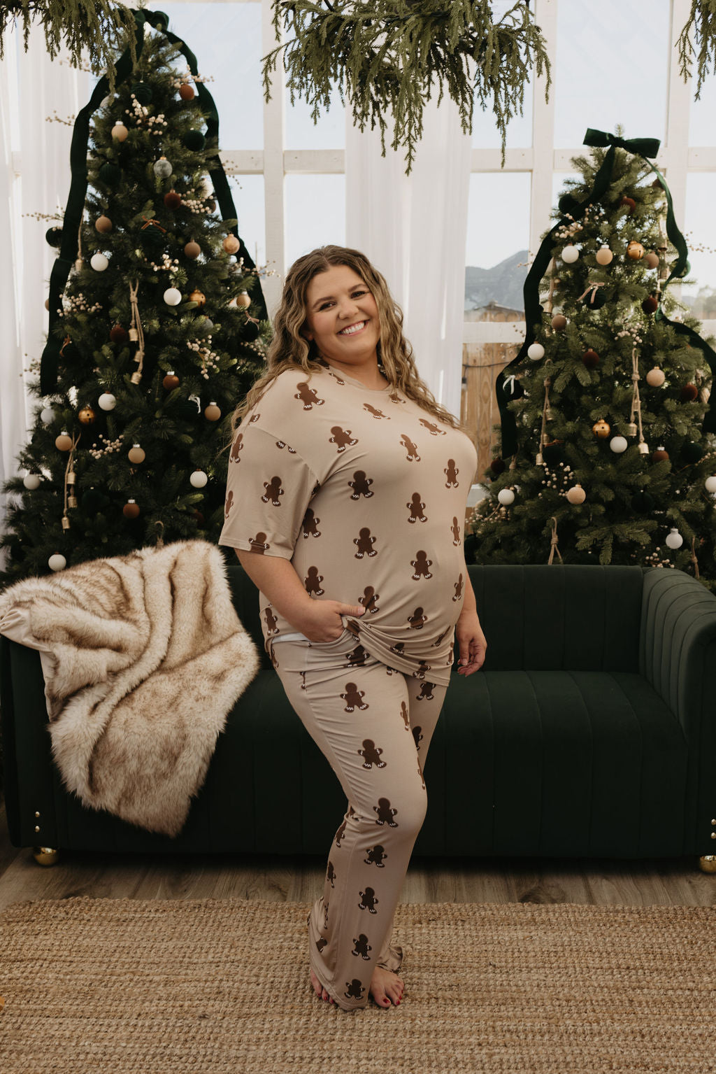 A person stands in front of two decorated Christmas trees, smiling as they wear the oversized Short Sleeve Women's Bamboo Pajamas with gingerbread prints by forever french baby. A dark green sofa draped with a fur blanket is in the background.