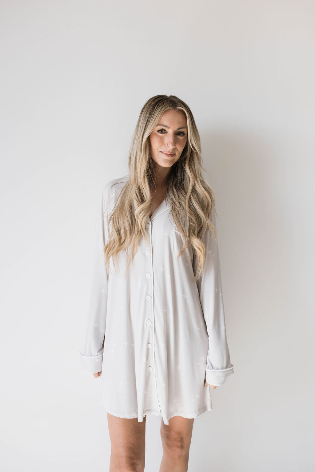 A woman with long, wavy blonde hair is posing against a plain white background. She is wearing an oversized, button-down Women's Bamboo Sleeping Dress by forever french baby and is looking directly at the camera with a slight smile.