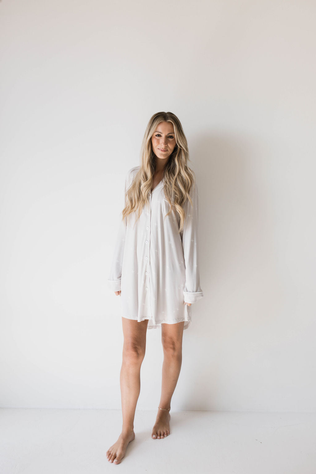 A woman with long, wavy blonde hair stands barefoot against a plain white background. She is wearing the Women's Bamboo Sleeping Dress by forever french baby, a white, long-sleeve button-up shirt dress made from eco-friendly and hypoallergenic materials, and smiling softly at the camera.