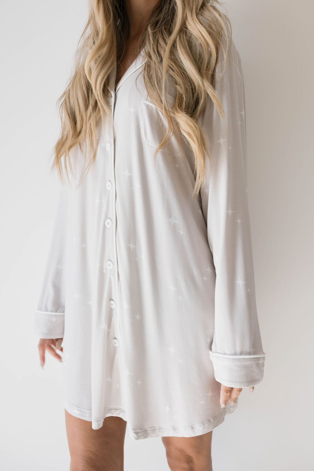 A woman with long, wavy blonde hair is wearing a light-colored, long-sleeved button-up Women's Bamboo Sleeping Dress | Sparkle! from forever french baby, adorned with subtle star patterns. The image is cropped above her knees, showcasing the upper half of her body and emphasizing the eco-friendly and hypo-allergenic fabric.