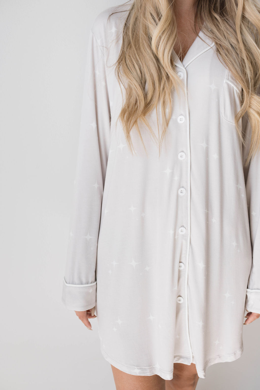 A person is wearing the forever french baby Women's Bamboo Sleeping Dress | Sparkle!, a loose-fitting, long-sleeved white night gown with buttons down the front. The hypo-allergenic nightshirt features a subtle star pattern. The person has long, wavy blond hair. The background is plain white, and only the lower face and body are visible in the image.