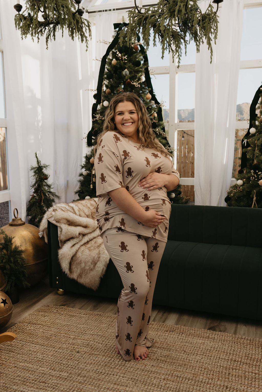 A smiling person stands indoors, wearing the Short Sleeve Women's Bamboo Pajamas in the Gingerbread pattern by forever french baby, gently holding their belly. Behind them is a green couch, a fur throw, and decorated Christmas trees, creating a festive atmosphere with hypo-allergenic clothing for ultimate comfort.