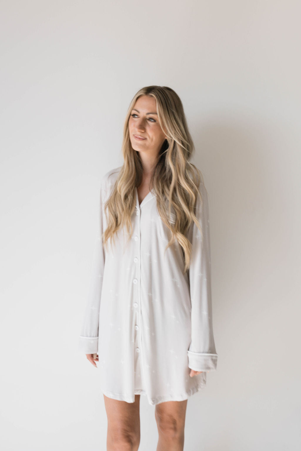 A person with long, wavy blonde hair stands against a plain white background. They are wearing an eco-friendly Women's Bamboo Sleeping Dress | Sparkle! from the brand forever french baby, featuring buttons down the front and sleeves rolled up to the elbows. The person is gazing slightly upward with a relaxed expression.