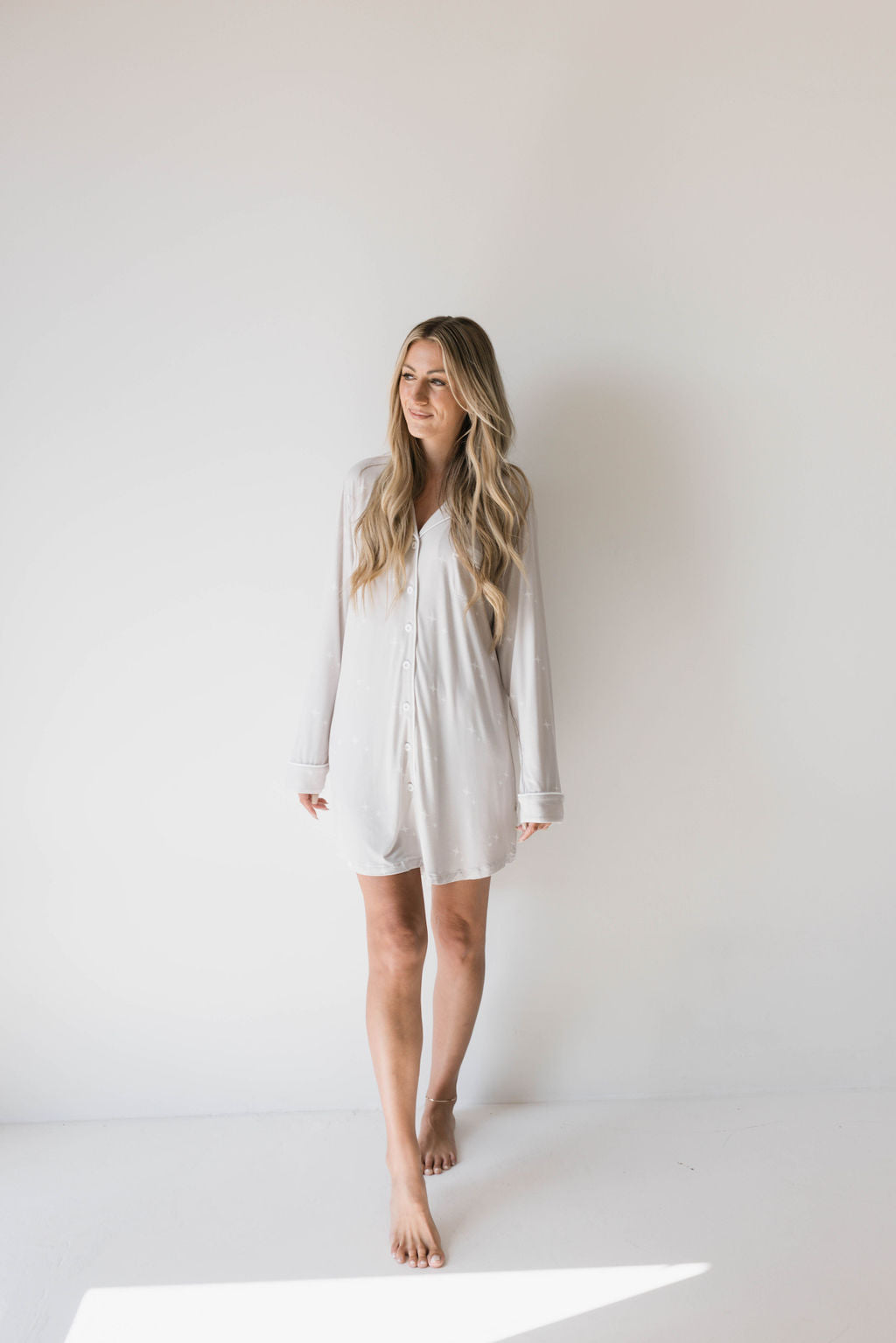 A person with long blonde hair stands barefoot against a plain white background, wearing a loose-fitting, eco-friendly "Women's Bamboo Sleeping Dress | Sparkle!" by forever french baby. They are smiling and looking slightly to the side. The overall mood is relaxed and casual.