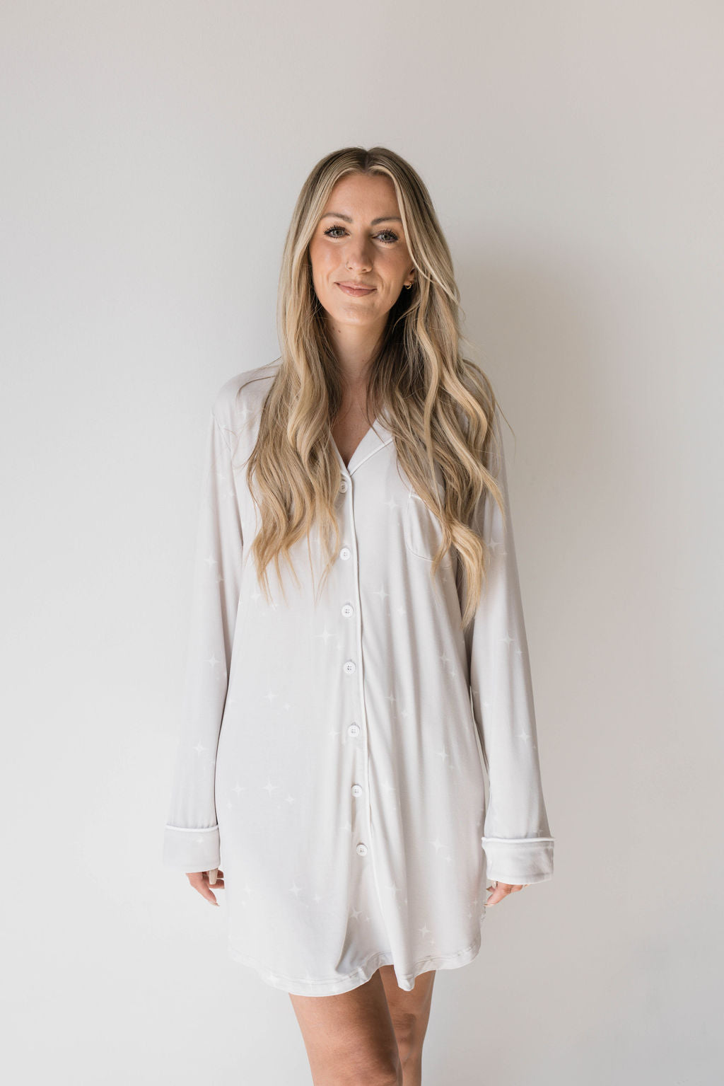 A woman with long, wavy blonde hair, wearing the eco-friendly Women's Bamboo Sleeping Dress by forever french baby in a light color, stands in front of a plain, light background. She has a subtle smile and her hands are relaxed by her sides.