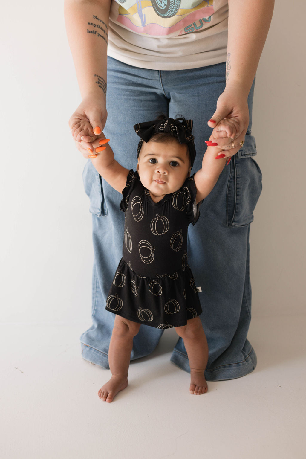 A baby girl in a Midnight Pumpkin Bamboo Ruffle Romper stands holding the hands of an adult behind her. The baby’s matching black bow complements her outfit perfectly. The adult, wearing a graphic t-shirt and loose blue jeans with bright orange nail polish, creates a charming scene fit for Forever French Baby memories.