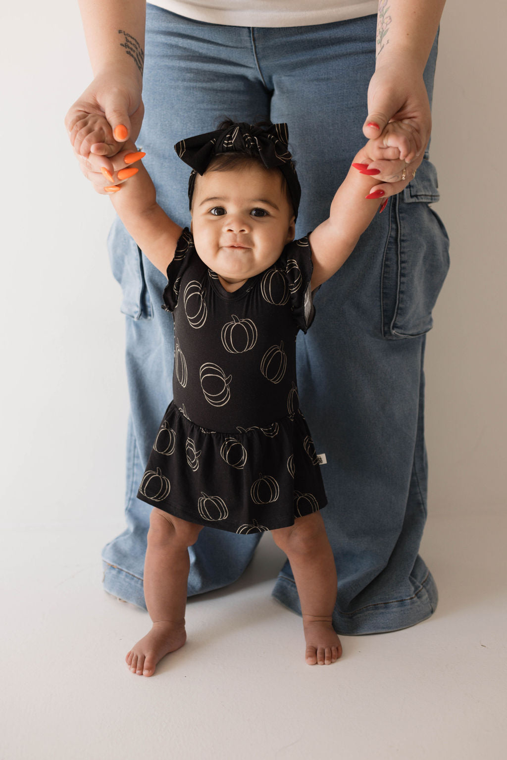 A cheerful baby wearing a Bamboo Ruffle Romper in Midnight Pumpkin, reminiscent of the forever french baby collection, is holding the hands of an adult in blue jeans. The baby stands with assistance, radiating joy. The adult's face remains out of frame.