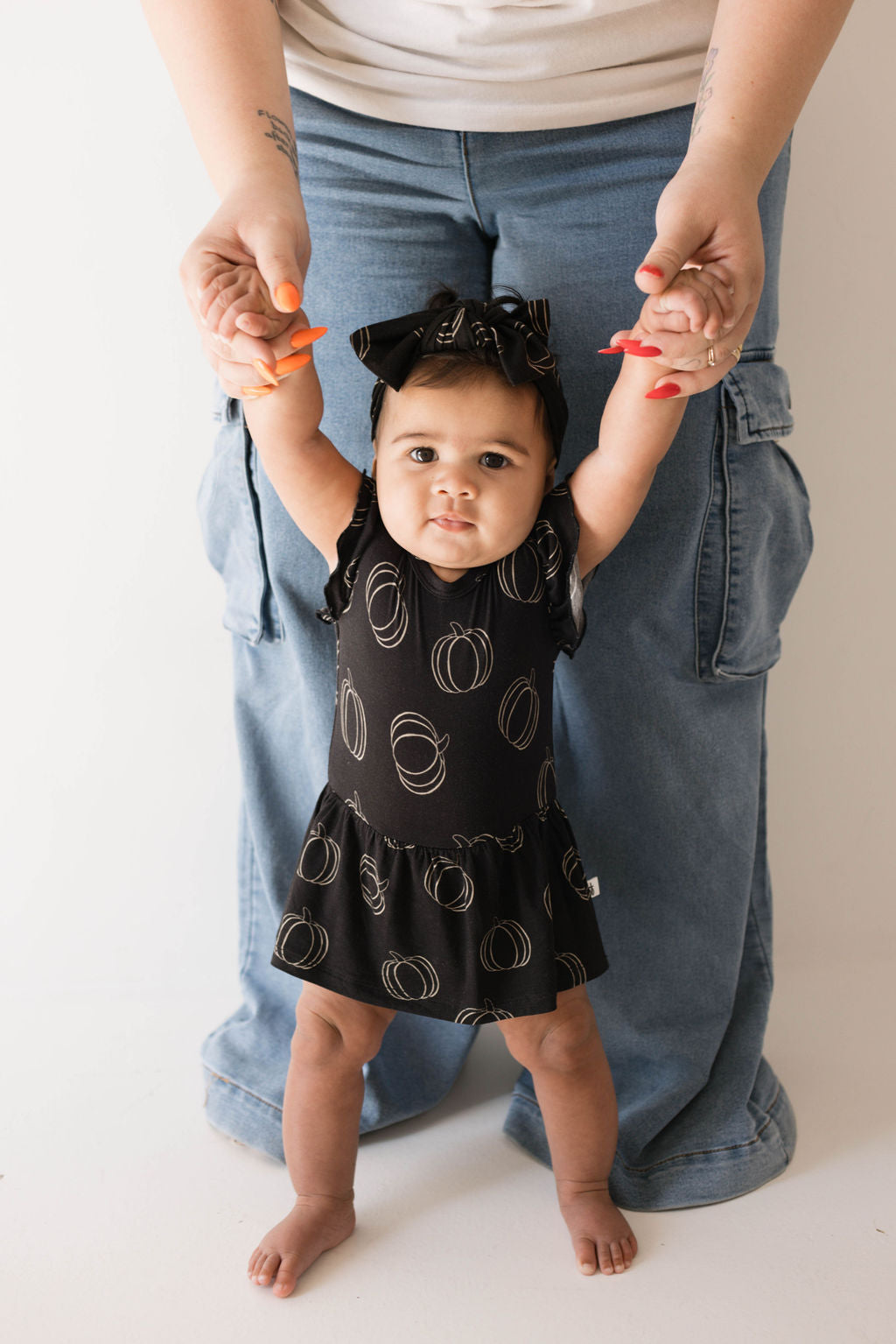 A baby dressed in the Forever French Baby's Bamboo Ruffle Romper in Midnight Pumpkin, featuring circular patterns and a matching headband, is being held up by an adult's hands. The adult, wearing a white shirt and blue jeans, supports the baby from behind by holding them up by their hands.