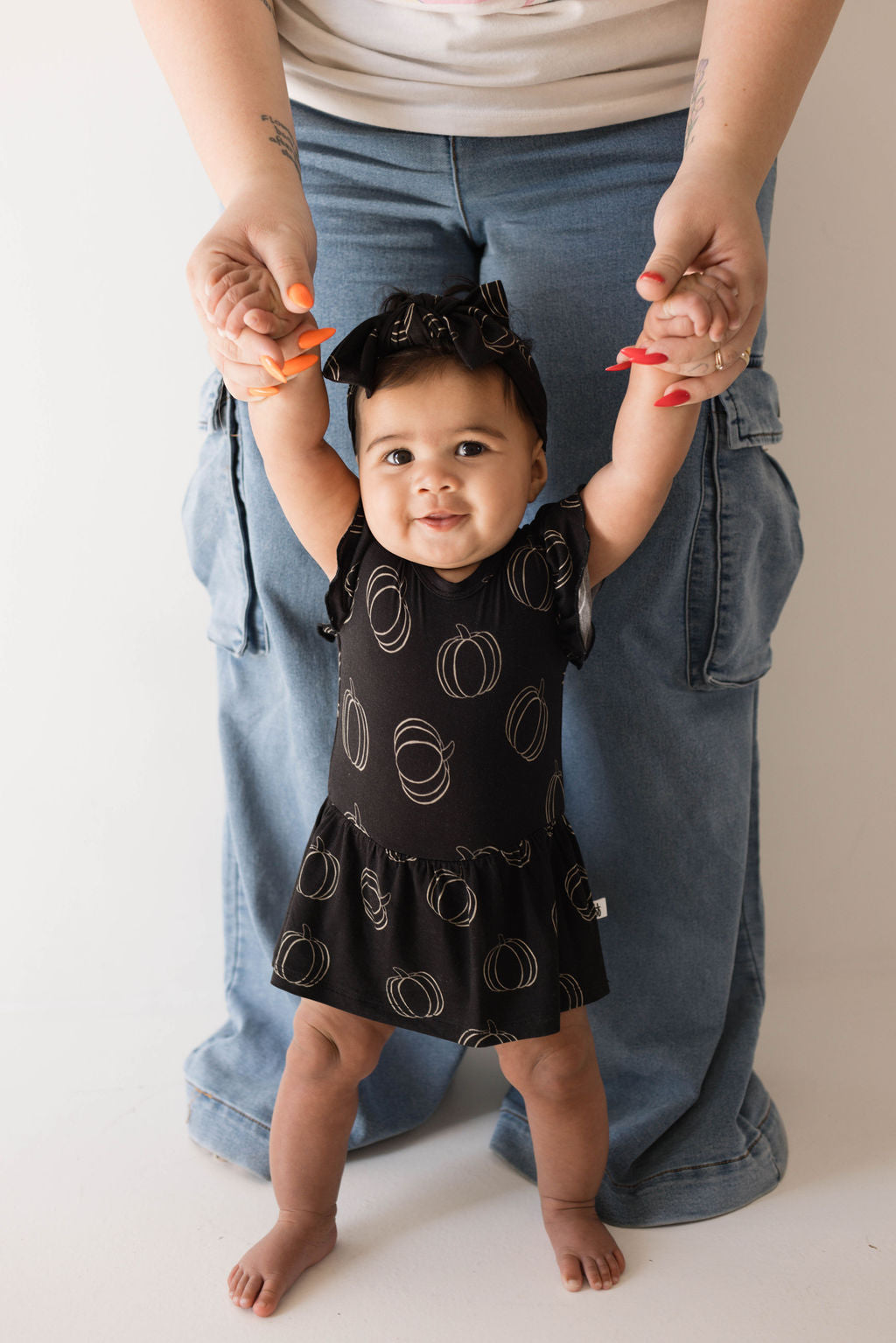 A toddler wearing a Bamboo Ruffle Romper | Midnight Pumpkin from forever french baby, along with a matching headband, is standing with the help of an adult behind, who is holding the toddler's hands. The outfit beautifully complements the adult's blue jeans and white shirt. The scene takes place indoors against a white background.