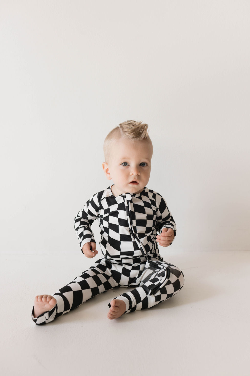 In front of a plain white background, a baby with light hair sits on the floor, wearing forever french baby Bamboo Zip Pajamas in Black & White Wavy Checkerboard. They appear to be looking slightly to the side and have a small crest of hair styled upward.