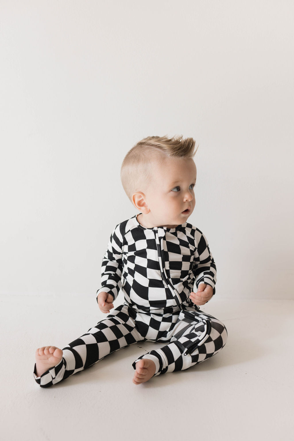 A baby with short blonde hair is sitting on the floor, wearing Forever French Baby's Bamboo Zip Pajamas in a Black & White Wavy Checkerboard pattern. The hypoallergenic clothing contrasts beautifully against the plain white background. The baby is looking slightly to the side, with a curious expression on their face.