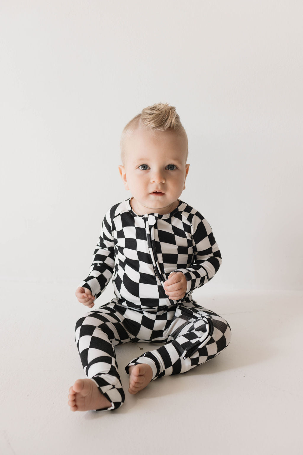 A baby is sitting on the floor wearing forever french baby's Bamboo Zip Pajamas in the Black & White Wavy Checkerboard pattern. The baby has light hair styled in a small fauxhawk, set against a plain, white background. The baby looks directly at the camera with a neutral expression, showcasing the comfort of hypo-allergenic clothing.