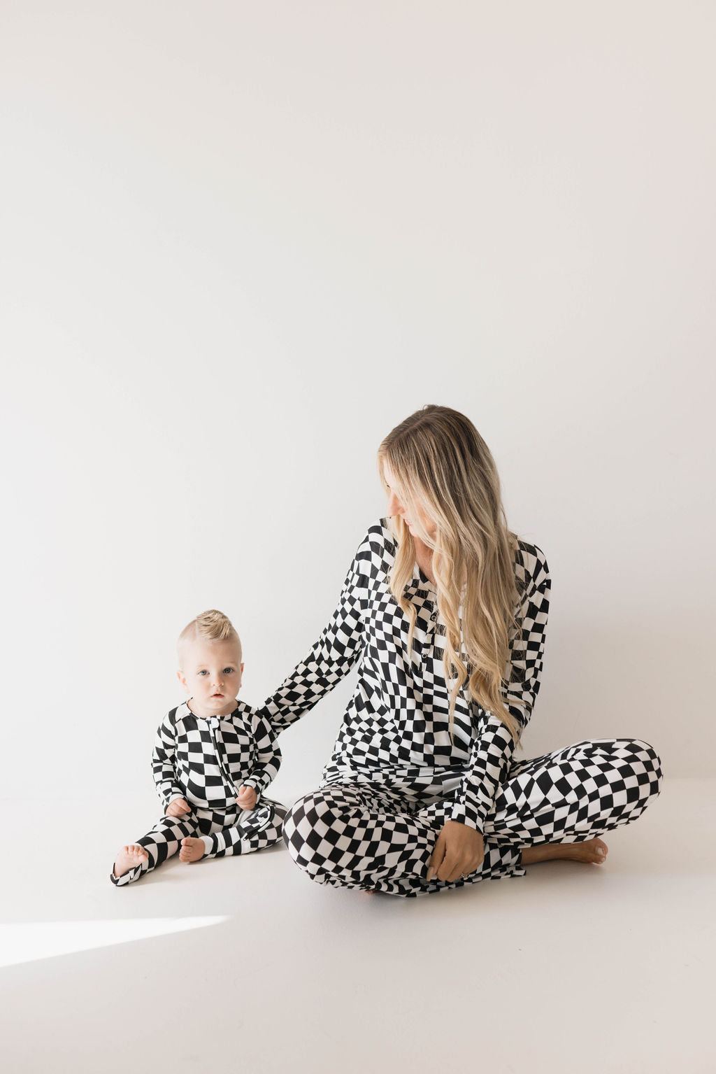 A woman with long blonde hair sits on the floor with her legs crossed, wearing Forever French Baby's Black & White Wavy Checkerboard Bamboo Zip Pajamas. Beside her is a young child in matching checkered baby sleepwear. They are both against a plain white background, and the woman smiles while looking at the child.