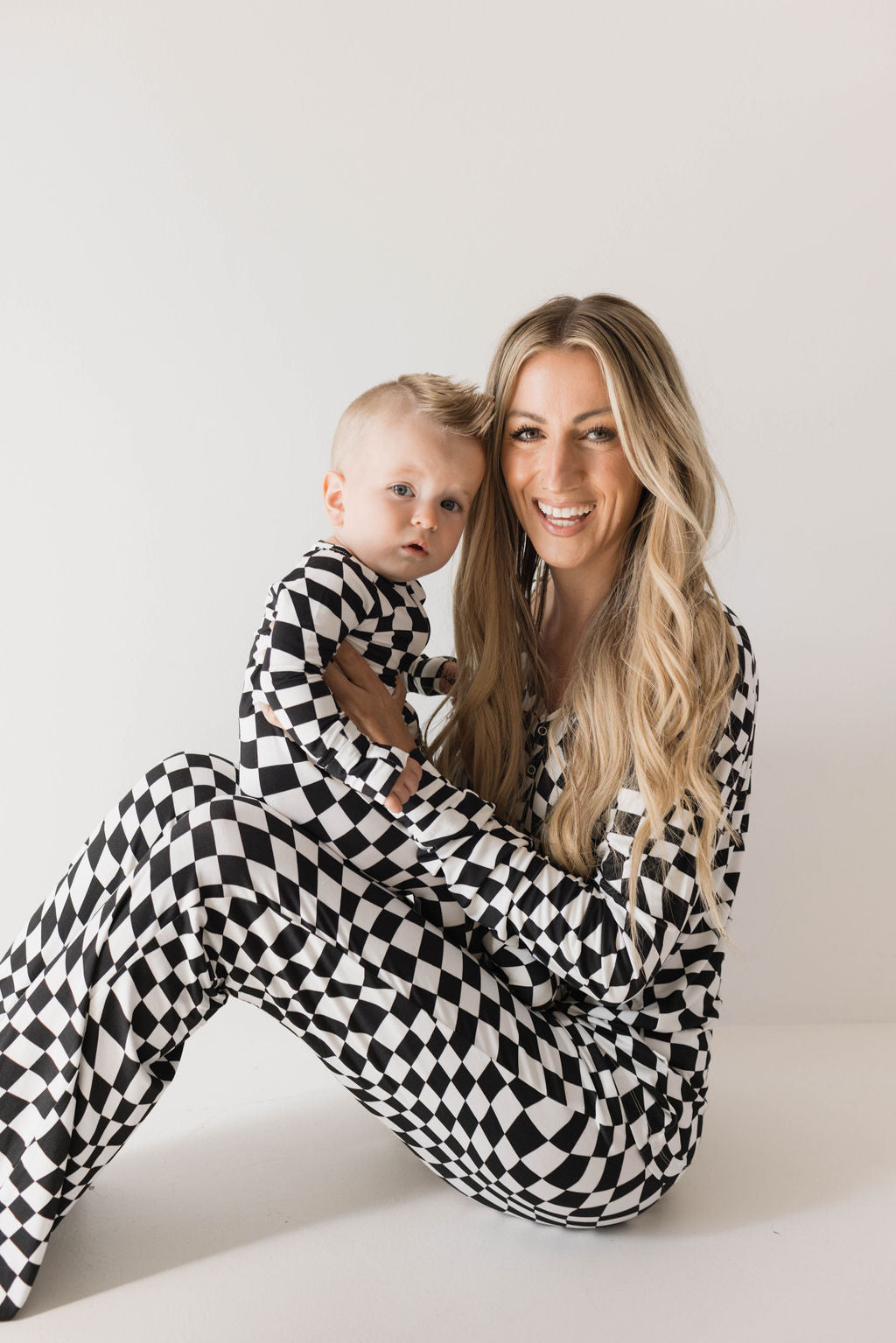 A woman with long blonde hair smiles as she sits on the floor, holding a baby. Both are dressed in matching hypo-allergenic Bamboo Zip Pajamas in Black & White Wavy Checkerboard print from forever french baby. The background is plain white.