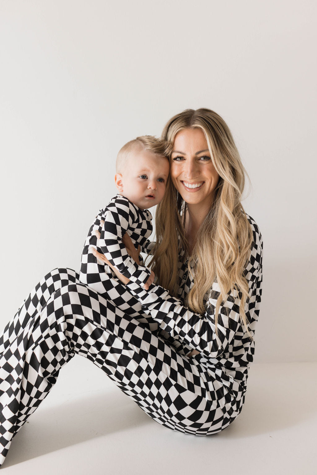 A woman with long, blonde hair and a smiling expression holds a baby. Both are dressed in matching "Bamboo Zip Pajamas | Black & White Wavy Checkerboard" from forever french baby. They sit against a plain white background, creating a clean and minimalistic look.