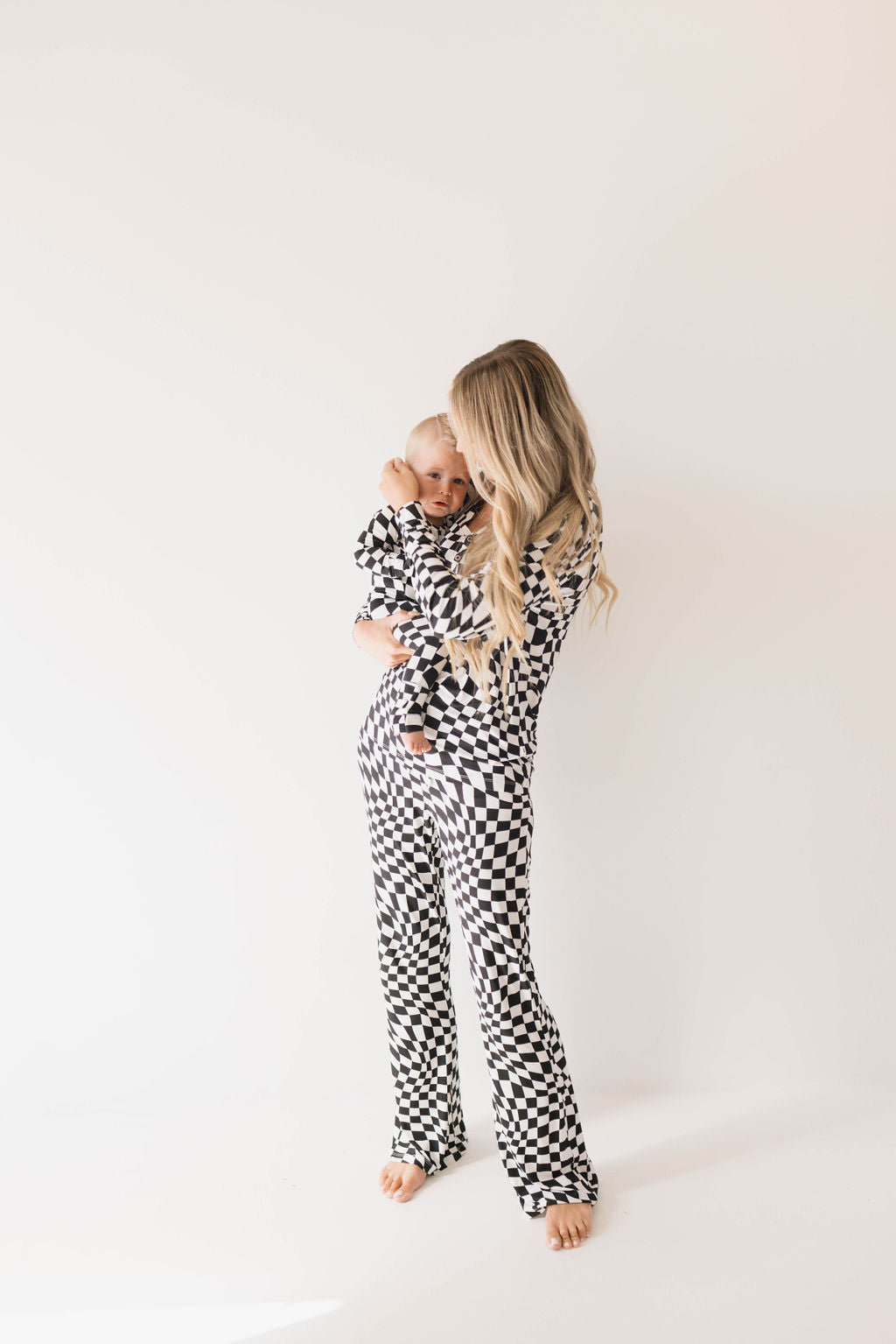 A woman with long blonde hair holds a baby in her arms, both dressed in matching "Bamboo Zip Pajamas | Black & White Wavy Checkerboard" by forever french baby. Standing against a plain white background, they look content and cozy.