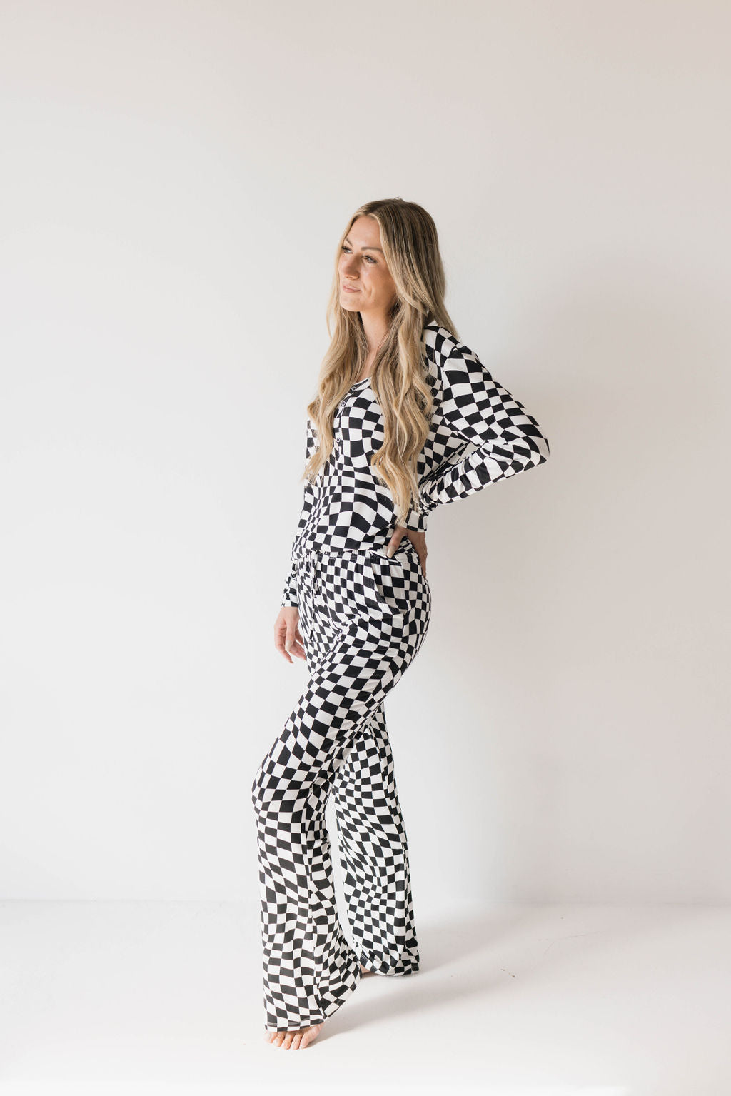 A woman with long blonde hair stands in front of a plain white background, wearing the Women's Bamboo Pajamas in Black & White Wavy Checkerboard from forever french baby. She is barefoot with her right hand resting on her hip, gazing slightly to the side with a relaxed and confident pose—perfect for breastfeeding moms.