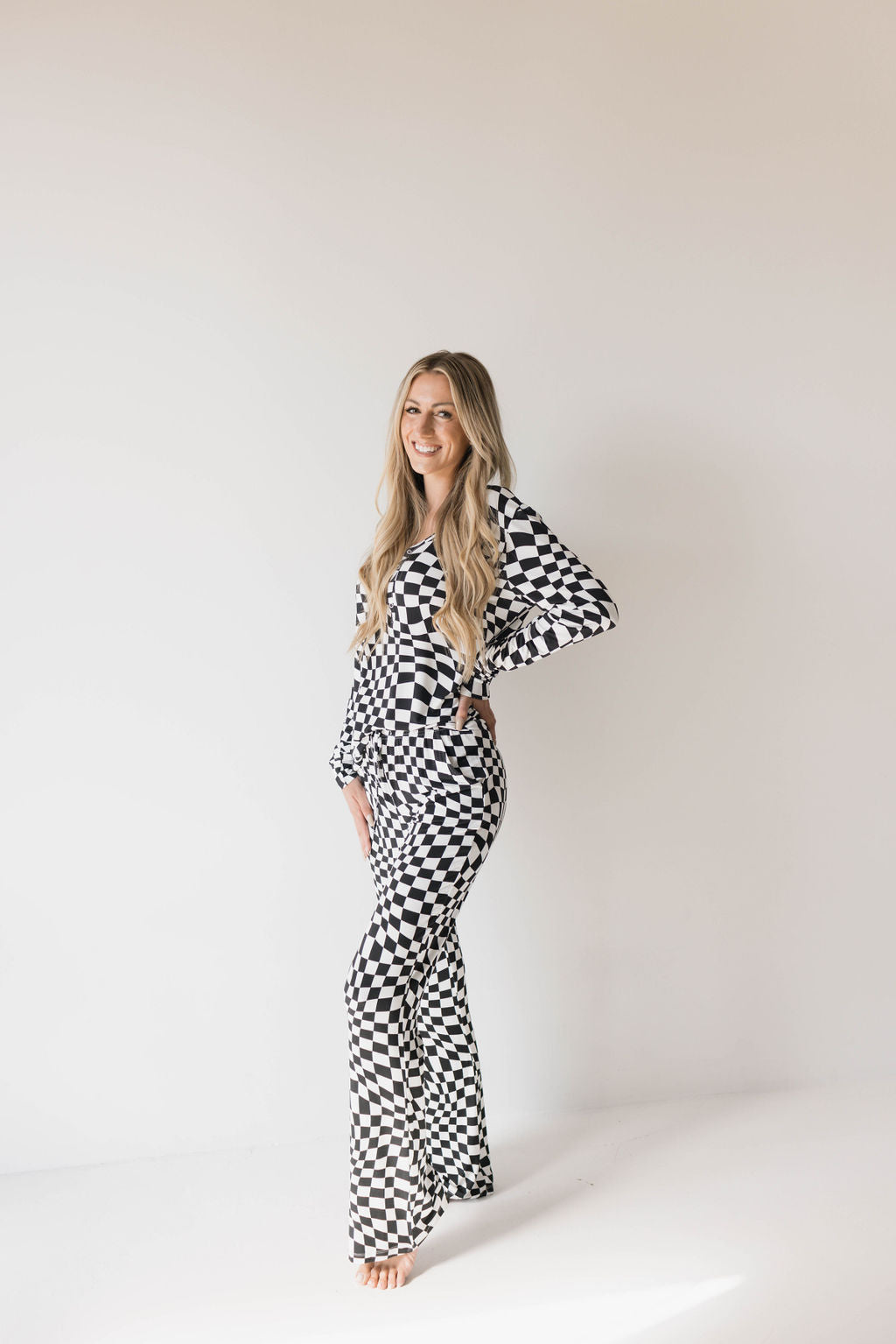 A person with long blonde hair stands against a plain white background, posing with one hand on their hip. They are wearing forever french baby's Women's Bamboo Pajamas in Black & White Wavy Checkerboard pattern, which includes a long-sleeve top and flared pants. Ideal for breastfeeding moms, they are barefoot and smiling.