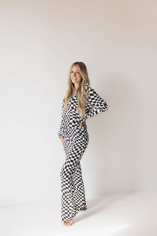 A person with long blonde hair stands against a plain white background, posing with one hand on their hip. They are wearing forever french baby's Women's Bamboo Pajamas in Black & White Wavy Checkerboard pattern, which includes a long-sleeve top and flared pants. Ideal for breastfeeding moms, they are barefoot and smiling.