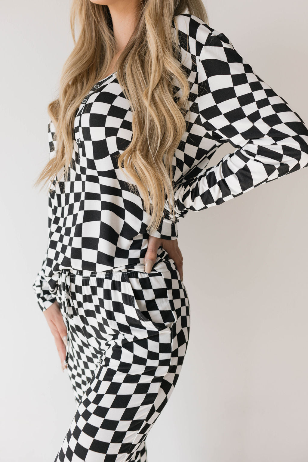 A person with long, wavy hair is wearing a matching black and white wavy checkerboard outfit from forever french baby, posing with one hand on their hip and the other by their side. The consistent pattern across the top and pants creates a cohesive look reminiscent of stylish Women's Bamboo Pajamas.