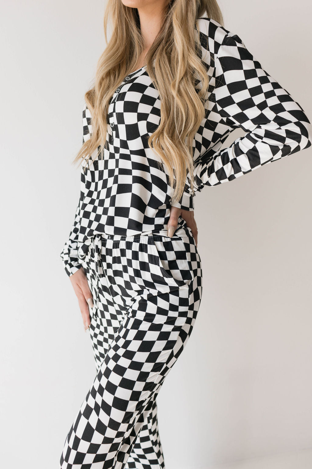 A woman with long blonde hair wears the Women's Bamboo Pajamas in Black & White Wavy Checkerboard by forever french baby. She stands with one hand on her hip, posing against a plain white background.