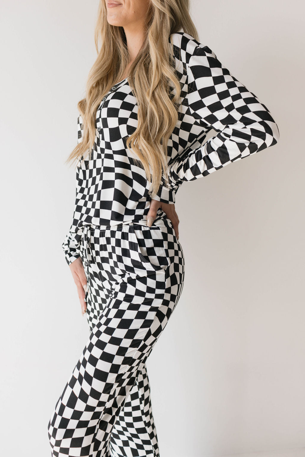 A person stands against a white background wearing the Women's Bamboo Pajamas in Black & White Wavy Checkerboard from forever french baby, featuring a long-sleeve top and matching pants. Their long blonde hair flows down past their shoulders, and their left hand rests on their hip.