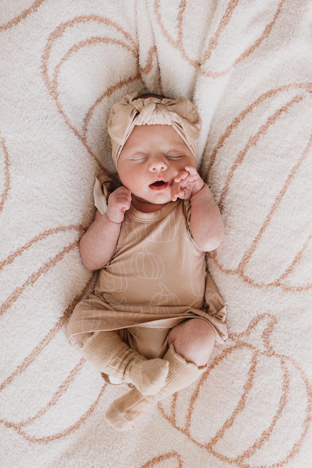 A newborn baby sleeps peacefully on a soft, textured blanket with an abstract pattern. The baby is dressed in a matching beige outfit, complete with an adjustable Bamboo Head Wrap from forever french baby. The onesie and socks feature a subtle pumpkin design, adding a cute touch to the scene.