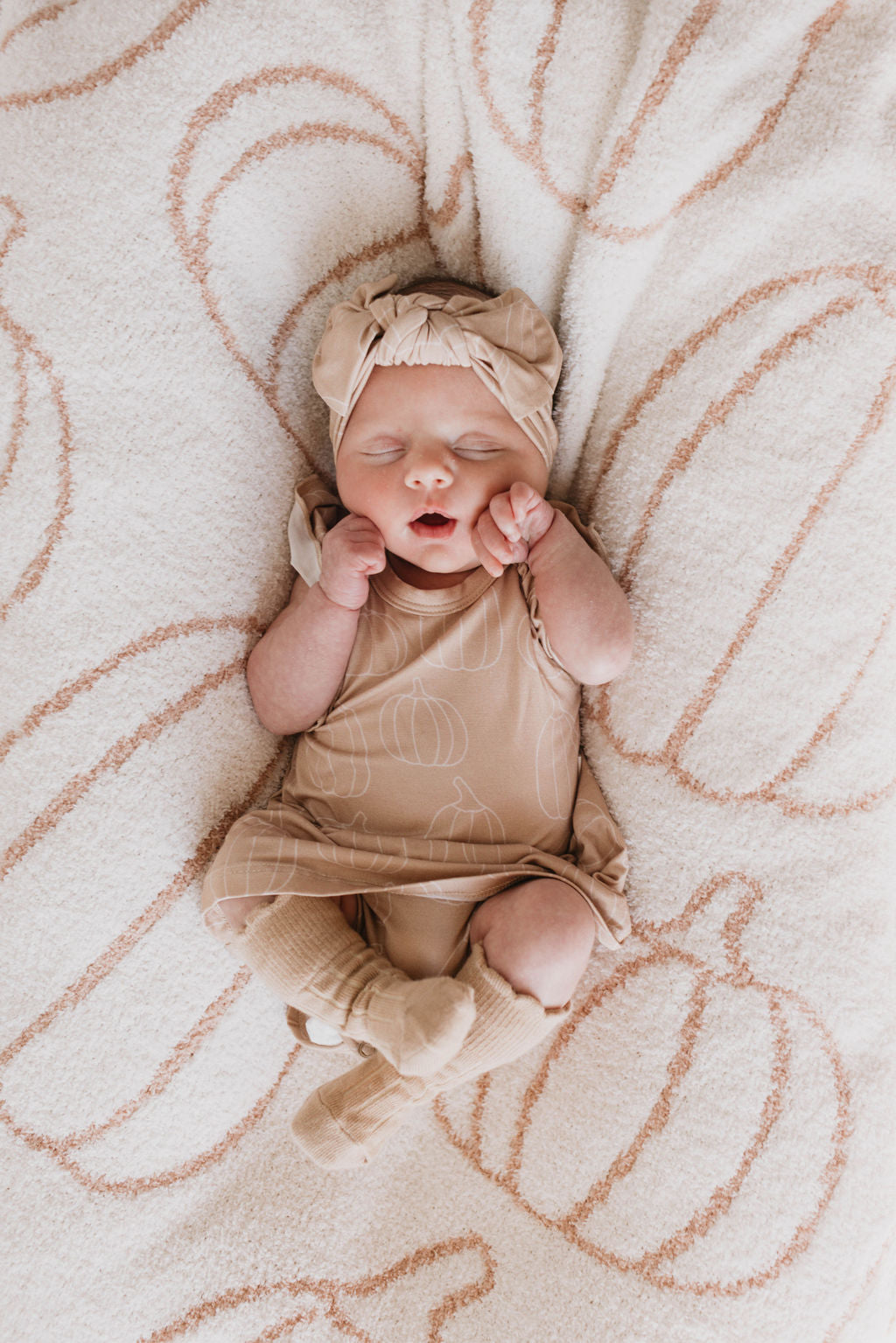 A newborn baby sleeps peacefully on a soft, white blanket with a pumpkin pattern. Dressed in the Bamboo Ruffle Romper in Pumpkin Pie from forever french baby, along with a matching hat and socks, the baby’s hands are near their face in a relaxed pose.