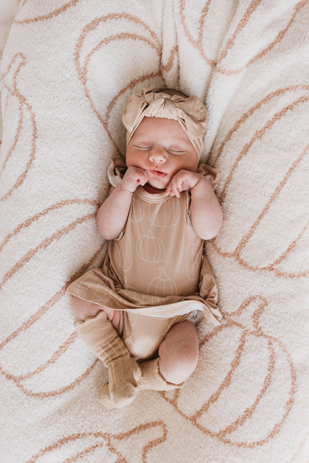 A newborn baby lies peacefully on a pumpkin-patterned blanket, dressed in a light brown outfit and an adjustable Bamboo Head Wrap | Pumpkin Pie by forever french baby. The baby has their tiny hands near their face and is wearing cozy socks, resting comfortably.