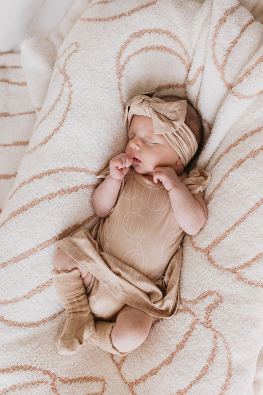 A sleeping baby adorned in a beige outfit featuring pumpkin patterns and an adjustable Bamboo Head Wrap | Pumpkin Pie from forever french baby lies on a soft, beige blanket with brown swirls. The baby's arms are slightly raised near its face, and one leg is playfully bent.