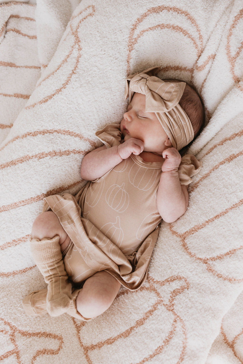 A newborn baby sleeps peacefully on a textured blanket, dressed in a light brown outfit with a pumpkin pattern, cozy socks, and an adjustable Bamboo Head Wrap | Pumpkin Pie by forever french baby adorned with a bow. The baby's hands rest near its face, creating an adorable and serene scene.