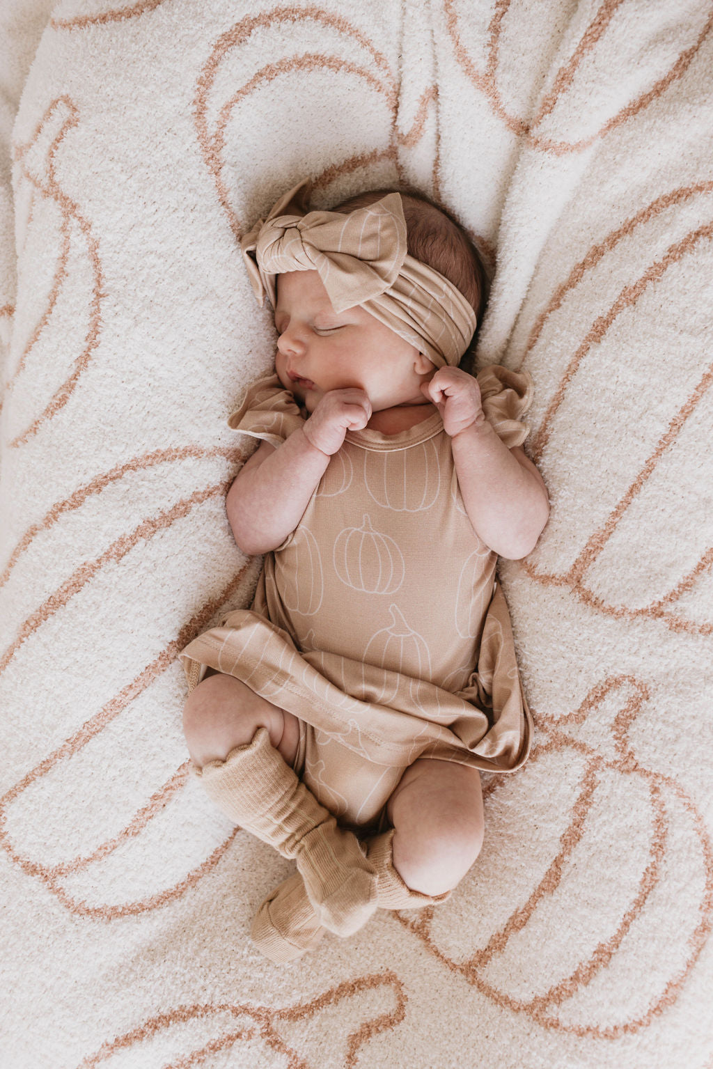 A newborn baby sleeps peacefully on a cozy, pumpkin-patterned blanket. The baby is dressed in a matching beige outfit featuring pumpkin designs, complete with the Bamboo Head Wrap | Pumpkin Pie from forever french baby, a dress, and socks. Their tiny fists are gently clenched near their face.