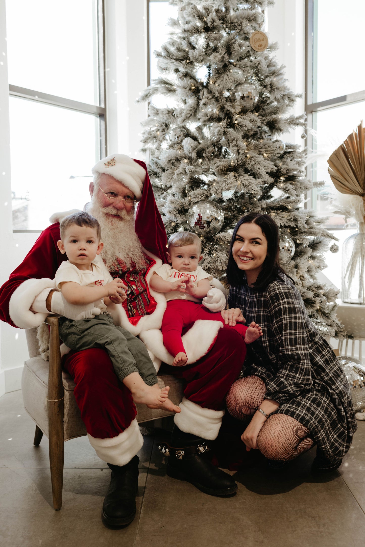 A person dressed as Santa Claus sits on a chair with two young children in their lap, each child perched on one knee. A woman kneels beside them, smiling warmly. They are in front of a beautifully decorated Christmas tree adorned with lights, all within a room featuring large windows—the perfect setting for heartwarming Santa Photos by Amanda Riley Photos' special edition: Santa Photos | Forever French Baby x Amanda Riley Photos.