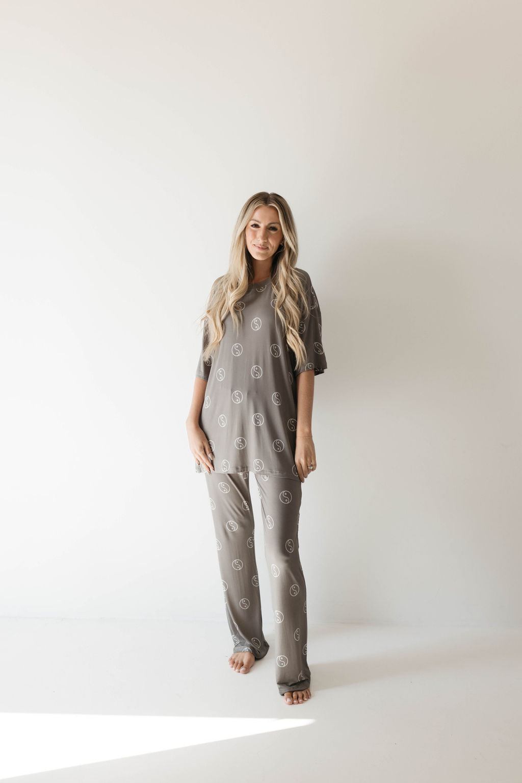 A woman with long blonde hair stands barefoot in a minimalist white space. She is wearing the Short Sleeve Women's Bamboo Pajamas in the Sage Yin Yang print by forever french baby. The relaxed and oversized pajama set, made from breathable fabric, complements her serene expression as she faces the camera with her hands by her sides.