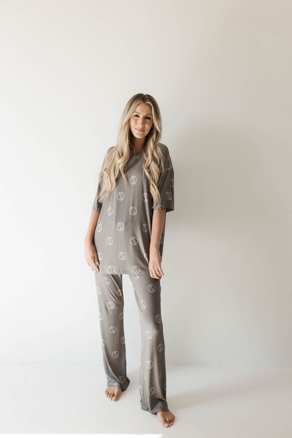 A person with long, blonde hair is standing against a plain white background. They are wearing an oversized pajama set from forever french baby made of breathable bamboo fabric, featuring a repetitive circular pattern called Sage Yin Yang. The loose-fitting, matching grey pajamas consist of a short-sleeved top and pants. They are barefoot and smiling gently at the camera.