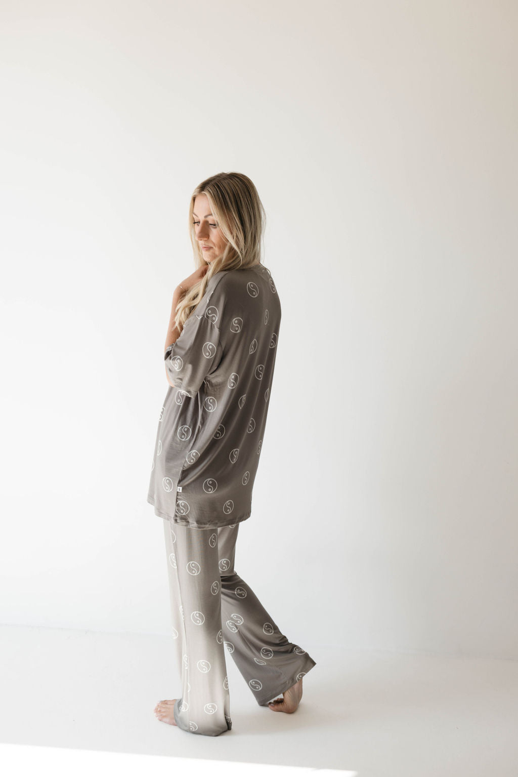 A woman with long hair wearing an oversized sage green pajama set from forever french baby, featuring a repeating pattern of white yin yang circles, poses against a plain white background. She faces to the side, with one hand touching her hair and the other resting at her side.