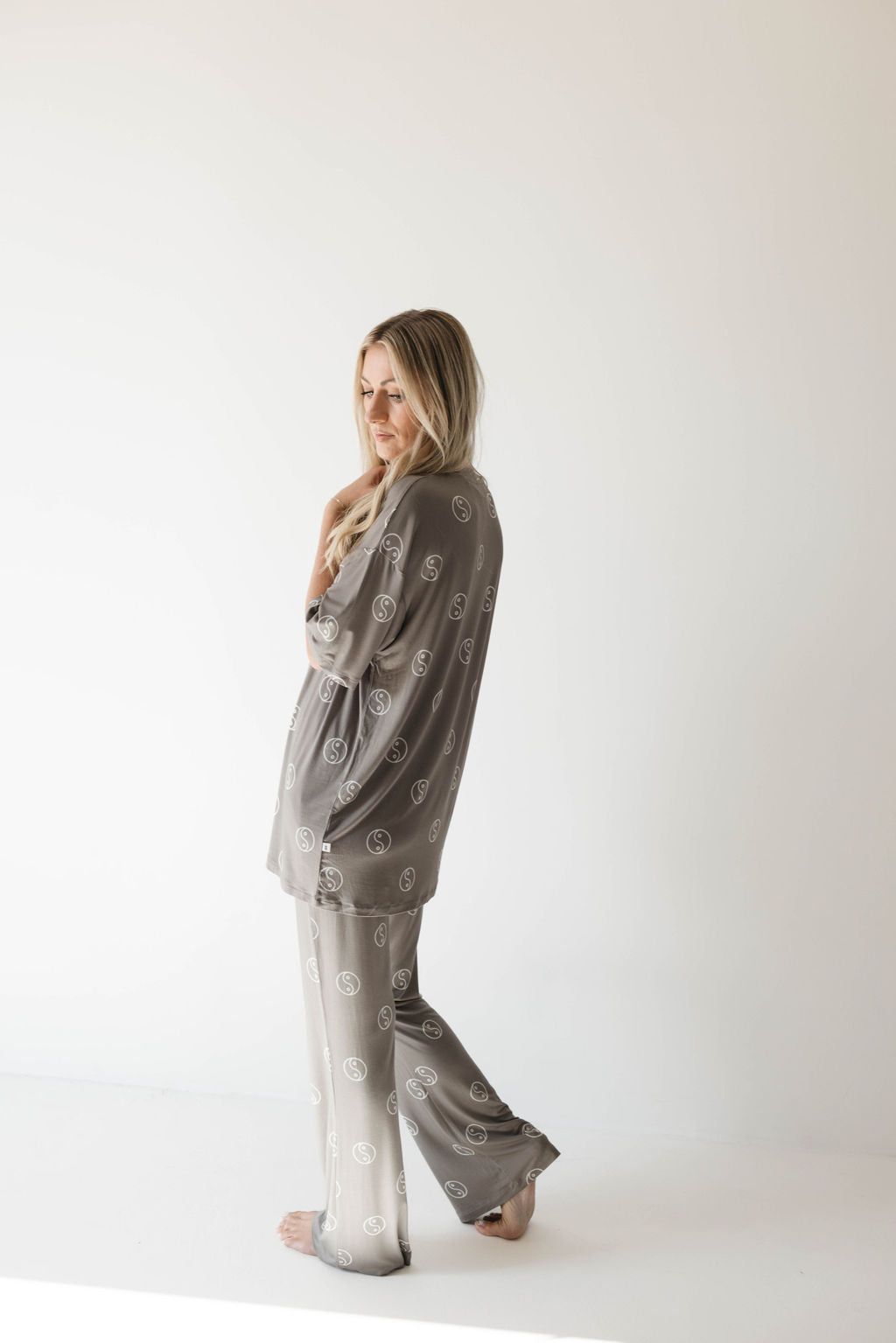 A person with long blonde hair stands barefoot against a white background. They are wearing an oversized, sage green bamboo pajama set featuring a repeating yin-yang pattern from forever french baby. One hand is resting near their neck, and they are looking off to the side with a calm expression.